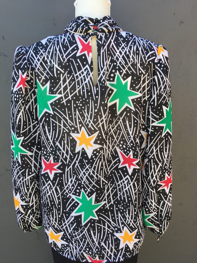 Puede incluir: Una blusa negra de cuello alto y mangas largas. La blusa tiene un estampado de estrellas blancas, verdes, rojas y amarillas sobre un fondo negro.