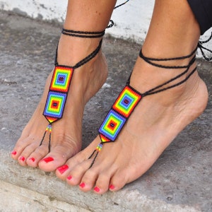 May include: A pair of rainbow beaded barefoot sandals with black cords. The sandals are tied around the ankles and have a beaded design with two squares.