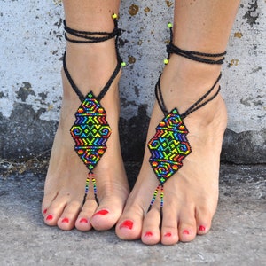 May include: A pair of colorful beaded barefoot sandals with black cords. The sandals feature a geometric pattern in shades of red, orange, yellow, green, blue, and purple.