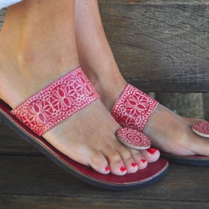 Könnte beinhalten: Rote Ledersandalen mit weißen Blumenstickereien und einem runden, roten Lederzehenring. Die Sandalen haben eine flache Sohle und werden an den Füßen einer Frau getragen.