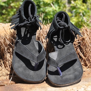 May include: A pair of black suede sandals with fringe detailing. The sandals have a thong style strap and a flat sole.