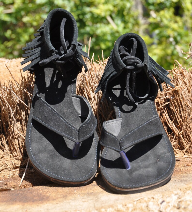 May include: A pair of black suede sandals with fringe detailing. The sandals have a thong style strap and a flat sole.