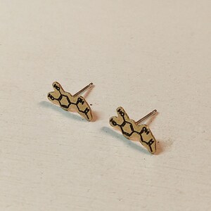 May include: A pair of gold-coloured stud earrings with a black chemical structure design. The earrings are small and delicate, with a post backing. The design is a repeating pattern of connected shapes.