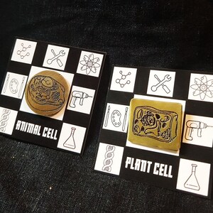 May include: Two science-themed pins. One is round and labeled "ANIMAL CELL," the other is square and labeled "PLANT CELL." Both are gold-colored and set on black and white checkered backgrounds with science-related icons.