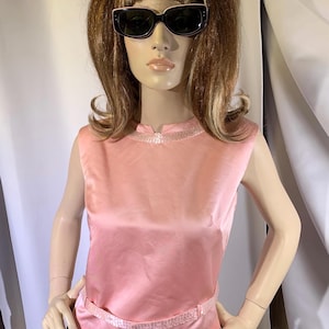 May include: A sleeveless, light pink satin blouse featuring a round neckline with subtle sequin detailing and a matching sequin-embellished belt at the waist. The top has a smooth, lustrous finish and a straight hemline, providing a clean, structured look.