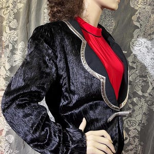 May include: A black velvet blazer with silver trim and a red undergarment. The blazer has a unique design with a curved collar and a silver button.