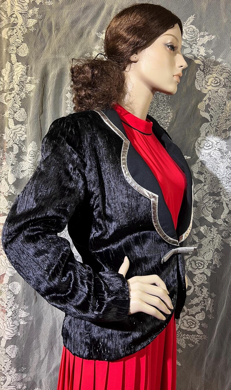 May include: A black velvet blazer with silver trim and a red undergarment. The blazer has a unique design with a curved collar and a silver button.