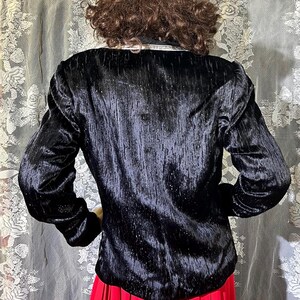 May include: A black velvet blazer with a sparkly texture. The blazer has long sleeves and a classic fit. The model is wearing a red pleated skirt.