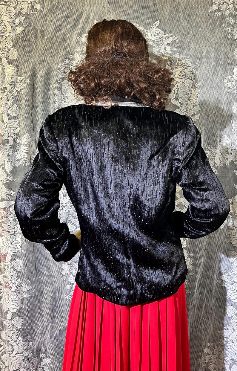 May include: A black velvet blazer with a sparkly texture. The blazer has long sleeves and a classic fit. The model is wearing a red pleated skirt.