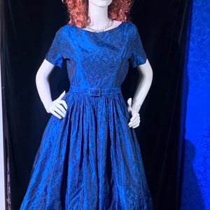May include: A vintage blue dress with a floral pattern. The dress features short sleeves, a fitted bodice, and a flared skirt. It has a matching belt and is displayed on a mannequin with a wig, sunglasses, and jewelry. The dress is a vibrant shade of blue.