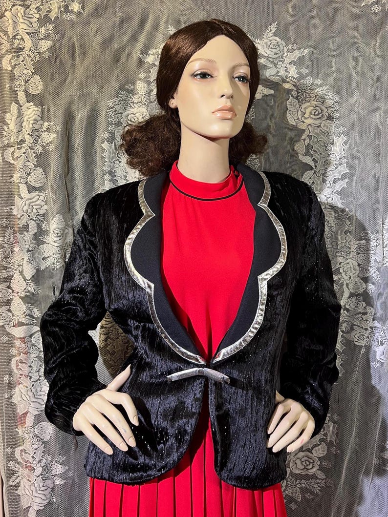 May include: A black velvet blazer with a silver trim and a decorative buckle closure. The blazer has a scalloped collar and a fitted silhouette.