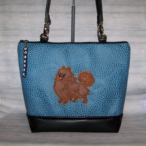 May include: A blue tote bag with a textured pattern and a black base. The bag features a brown Pomeranian dog applique. The bag has black handles and a zipper closure with a paw print charm.