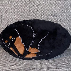 May include: A black and brown dachshund dog-shaped cushion. The cushion is oval and features a sleeping dog design with white outline details. The dog's face and paws are brown, and the body is black.