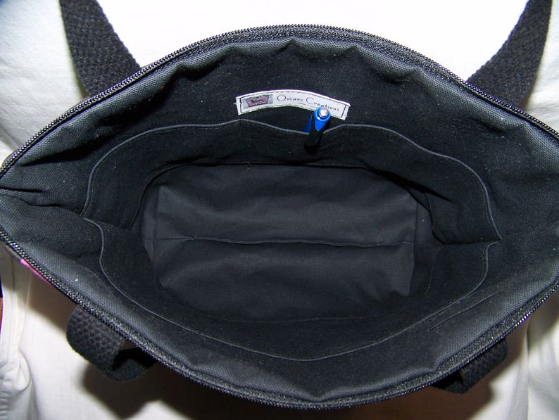 May include: Open view of a black tote bag with a zipper closure and black handles. The interior is black with multiple pockets and a pen. The bag has a label that reads "Oceans Creations".