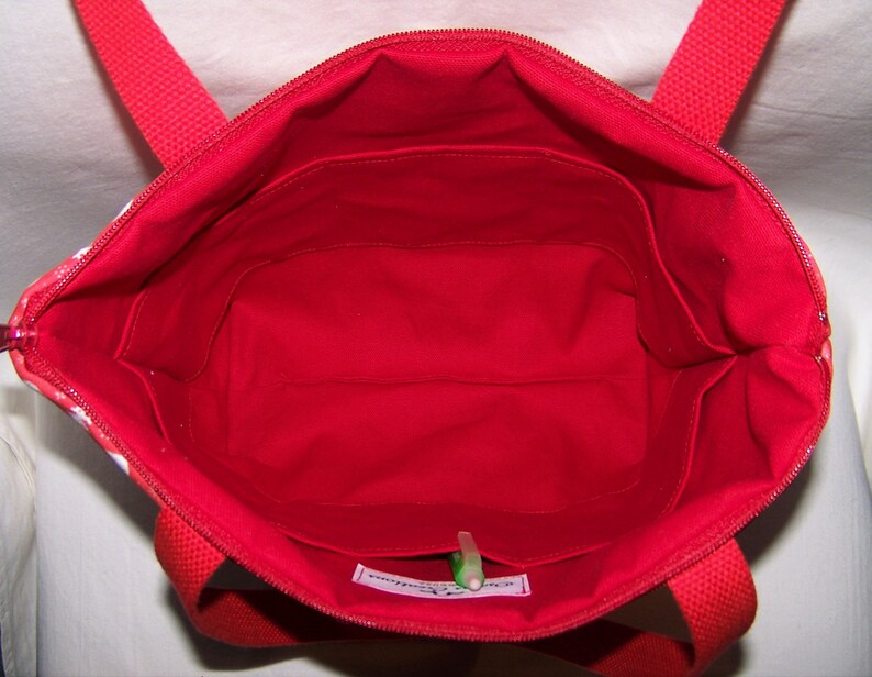 Pu&ograve; includere: Vista interna di una borsa tote rossa con chiusura a zip e manici rossi. L'interno &egrave; rosso vivo con pi&ugrave; tasche. Il tessuto esterno presenta un motivo floreale bianco. Si vede una penna verde.