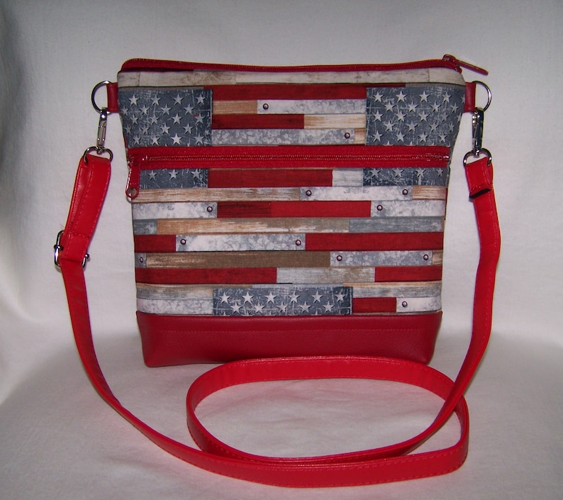 Pu&ograve; includere: Borsa a tracolla rettangolare con base e cinturino in pelle rossa. Il corpo principale della borsa presenta una stampa che ricorda la bandiera americana, con strisce rosse e bianche e un campo blu di stelle. Cerniere e hardware argentati completano il design.