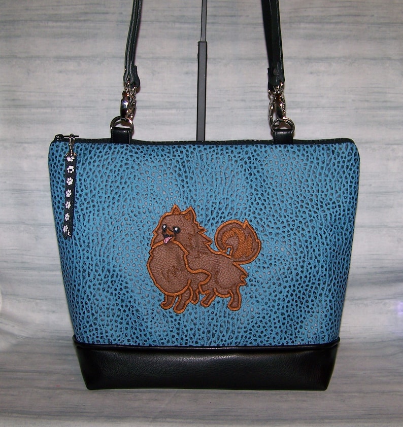 May include: A blue tote bag with a textured pattern and a black base. The bag features a brown Pomeranian dog applique. The bag has black handles and a zipper closure with a paw print charm.