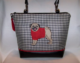 May include: A black and white houndstooth tote bag with a black leather bottom. The bag features an embroidered image of a pug wearing a red sweater.