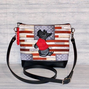 Pu&ograve; includere: Borsa a tracolla con motivo a bandiera americana, base e cinturino in pelle nera. La borsa ha una chiusura a zip e un ciondolo. Un cane dei cartoni animati ricamato con una camicia rossa &egrave; sul davanti.