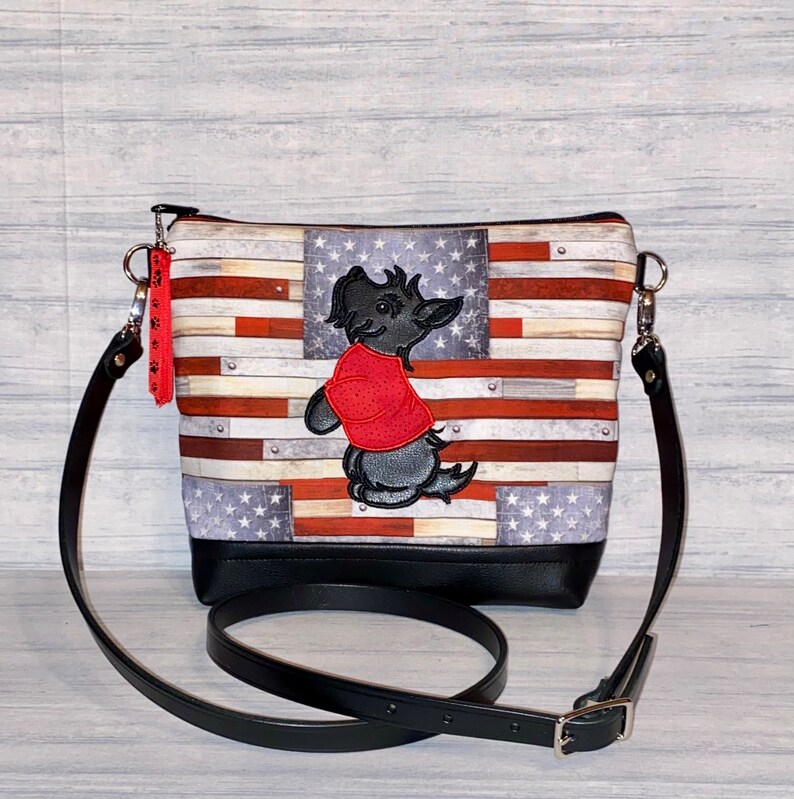 Pu&ograve; includere: Borsa a tracolla con motivo a bandiera americana, base e cinturino in pelle nera. La borsa ha una chiusura a zip e un ciondolo. Un cane dei cartoni animati ricamato con una camicia rossa &egrave; sul davanti.