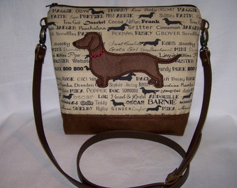 May include: A brown leather crossbody bag with a beige fabric front panel. The fabric panel features a dachshund silhouette with red beads around its neck. The fabric is covered in a pattern of black dachshund silhouettes and names, including "Maggie", "Faith", "Pretzel", "Missy Annie", "Thorny", "Love", "Pinkie", "Cricket", "Skittles", "True Grit", "Daschel", "Cocoa", "Nathan", "Frank", "Gritter", "Cinnamon", "Jaeger", "Herman Husky", "Grover Scruffles", "Sweet Caroline", "Greta Girl", "Von Goorheart", "Kons", "Scooby", "Mini", "Dunder", "Killion", "Piper", "Buster", "Webster", "Duke", "Boo", "Rose", "Eiki", "Vie", "Ramba", "Dresden", "Pepper", "Doc", "Schnoodle", "Sophie", "Jazz", "Bentley", "Line", "Rambe", "Semgrean", "Dixie", "Oscar", "Lou", "Hansel", "Gretal", "Andouille", "Barnie", "Wiggle", "Ma", "Shelby", "Kylie", "Ginger", "Layla", "Mike", "Ma".