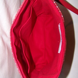 Pu&ograve; includere: Vista aperta di una pochette rossa con cerniera, interno rosso e piccole tasche. L'esterno &egrave; rosso con pois bianchi. Un cinturino rosso &egrave; attaccato alla cerniera.