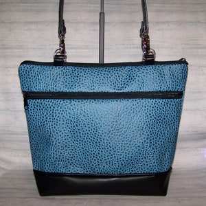 May include: A turquoise tote bag with a textured pattern and black accents. The bag features a black base, two zippered pockets, and black leather straps with silver hardware. The bag is designed with a structured shape.