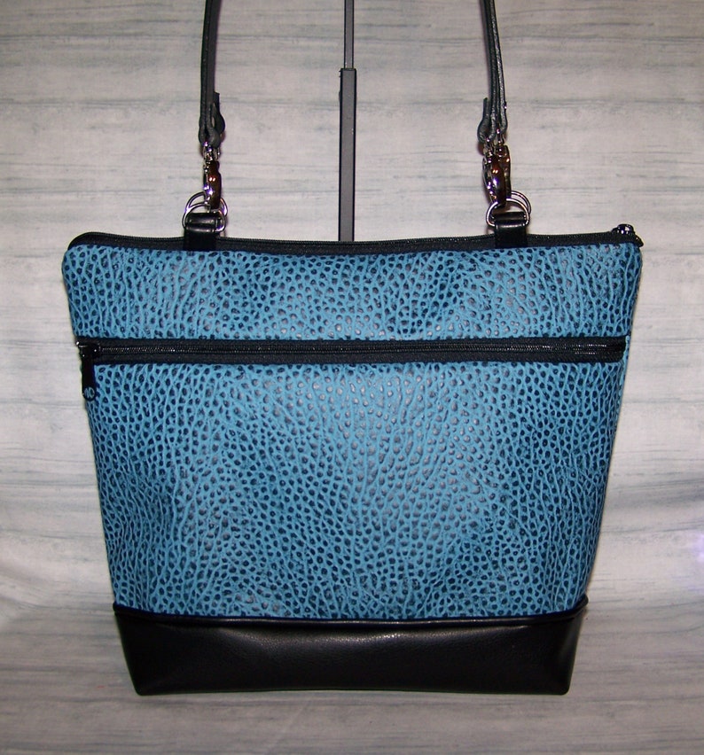 May include: A turquoise tote bag with a textured pattern and black accents. The bag features a black base, two zippered pockets, and black leather straps with silver hardware. The bag is designed with a structured shape.