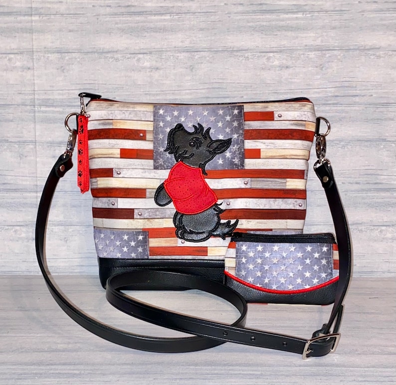Pu&ograve; includere: Una borsa a tracolla e un portafoglio abbinato con un design ispirato alla bandiera americana. La borsa ha un cinturino e una base in pelle nera, con un cane ricamato con una camicia rossa. Il portafoglio ha un bordo rosso.