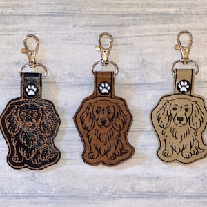 May include: Three dog-shaped keychains with gold-colored clasps. The keychains are brown, tan, and black, each featuring a different color scheme and a paw print detail. The dog designs are embroidered with detailed outlines.