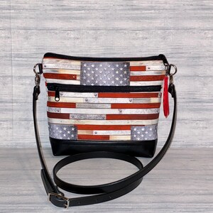 Pu&ograve; includere: Una borsa a tracolla con un design a bandiera americana. La borsa presenta un fondo nero, una tracolla regolabile nera e un nappa rossa. Il design della bandiera incorpora strisce rosse e bianche con un campo blu di stelle bianche.