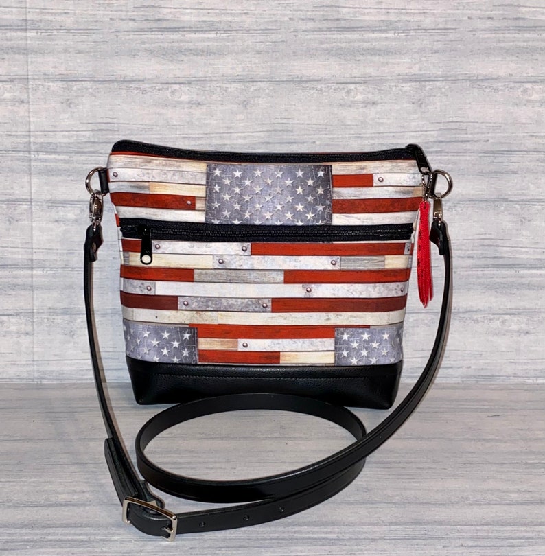 Pu&ograve; includere: Una borsa a tracolla con un design a bandiera americana. La borsa presenta un fondo nero, una tracolla regolabile nera e un nappa rossa. Il design della bandiera incorpora strisce rosse e bianche con un campo blu di stelle bianche.
