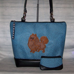 May include: A blue tote bag and matching coin purse with a textured pattern and a black bottom. The tote features an embroidered brown Pomeranian dog. The bag has black handles and a paw print zipper pull.