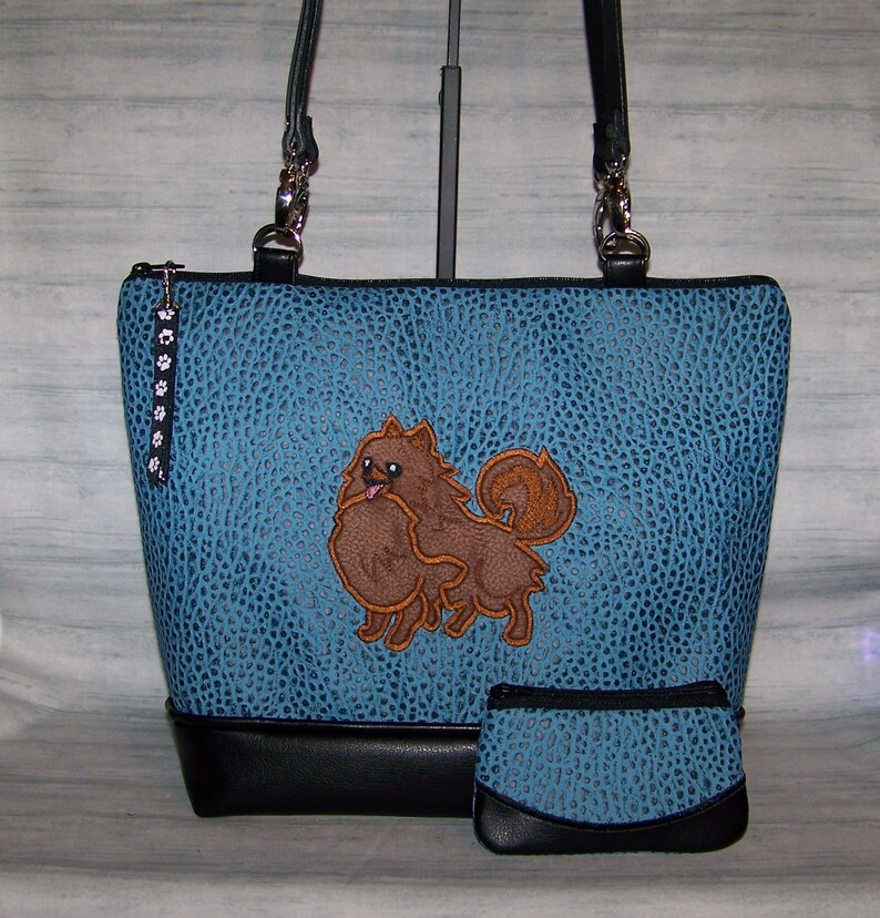 May include: A blue tote bag and matching coin purse with a textured pattern and a black bottom. The tote features an embroidered brown Pomeranian dog. The bag has black handles and a paw print zipper pull.