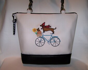 May include: White tote bag with a black base. The bag features an embroidered design of a dachshund wearing a red hat and scarf riding a blue bicycle with a basket of sunflowers and a pumpkin.