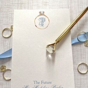 May include: A white notepad with a blue inscription that reads "The Future Mrs. Madeline Parker". The notepad has a gold ring with a diamond and a letter "P" in the center.