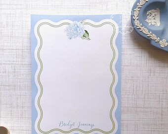 Può includere: Blocco note di cancelleria blu con un bordo verde e un'illustrazione ad acquerello di un'ortensia blu. Il blocco note ha il nome "Bridget Jennings" stampato in basso.