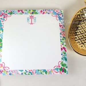 May include: A white notepad with a colorful floral border and the monogram "MTH" in pink and blue. The notepad is on a white surface next to a gold pineapple-shaped dish.