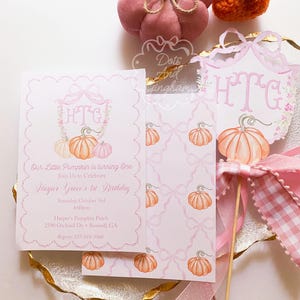 May include: Pink and white invitation suite with pumpkin illustrations. The invitation reads "Our Little Pumpkin is turning One" and includes event details. A decorative pumpkin-themed cake topper and decorative pumpkins are also present.