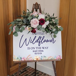 May include: A white sign with a floral border and the text "A Little Wildflower is on the way Baby Piper 02/13/24" in navy blue lettering.