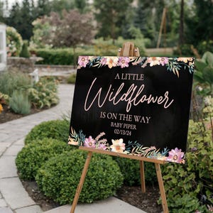 May include: A black and white floral baby shower sign with the text "A Little Wildflower is on the way" in pink script. The sign also includes the text "Baby Piper 02/13/24".