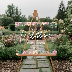 May include: A clear acrylic sign with a floral design and the text "A Little Wildflower is on the way Baby Piper 02/13/24".
