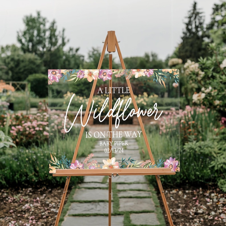 May include: A clear acrylic sign with a floral design and the text "A Little Wildflower is on the way Baby Piper 02/13/24".