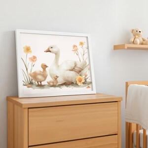 May include: A framed watercolor print featuring a white goose with two goslings amidst yellow and peach flowers. The artwork is displayed on a wooden dresser, creating a soft, inviting aesthetic. A teddy bear sits on a shelf in the background.