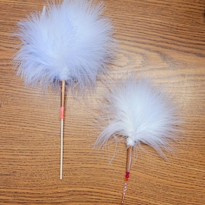 May include: Two white feather wands. The larger wand features a copper-colored metal section, a bright pink band, and a wooden handle. The smaller wand includes a copper-colored metal section, pink and orange beads, and a wire loop at the end.