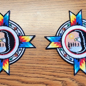 May include: Two embroidered patches with a Native American design. Each patch features a central circle with an eagle and trees, surrounded by a cross-shaped design in blue, yellow, and red. The patches are on a wooden surface.