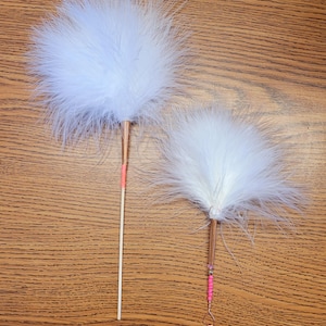 May include: Two white feather wands with copper-coloured metal ferrules. The left wand features a long, thin, light-coloured wooden handle with a small pink band. The right wand has a shorter handle decorated with pink beads and a wire loop at the end.