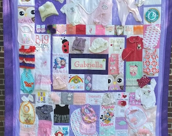 Puede incluir: Un edredón de bebé hecho a mano en tonos morados, con varias prendas y accesorios para bebés. El edredón incluye bodies, baberos, sombreros y una etiqueta con el nombre "Gabriella". El diseño es un patchwork con colores y patrones variados.