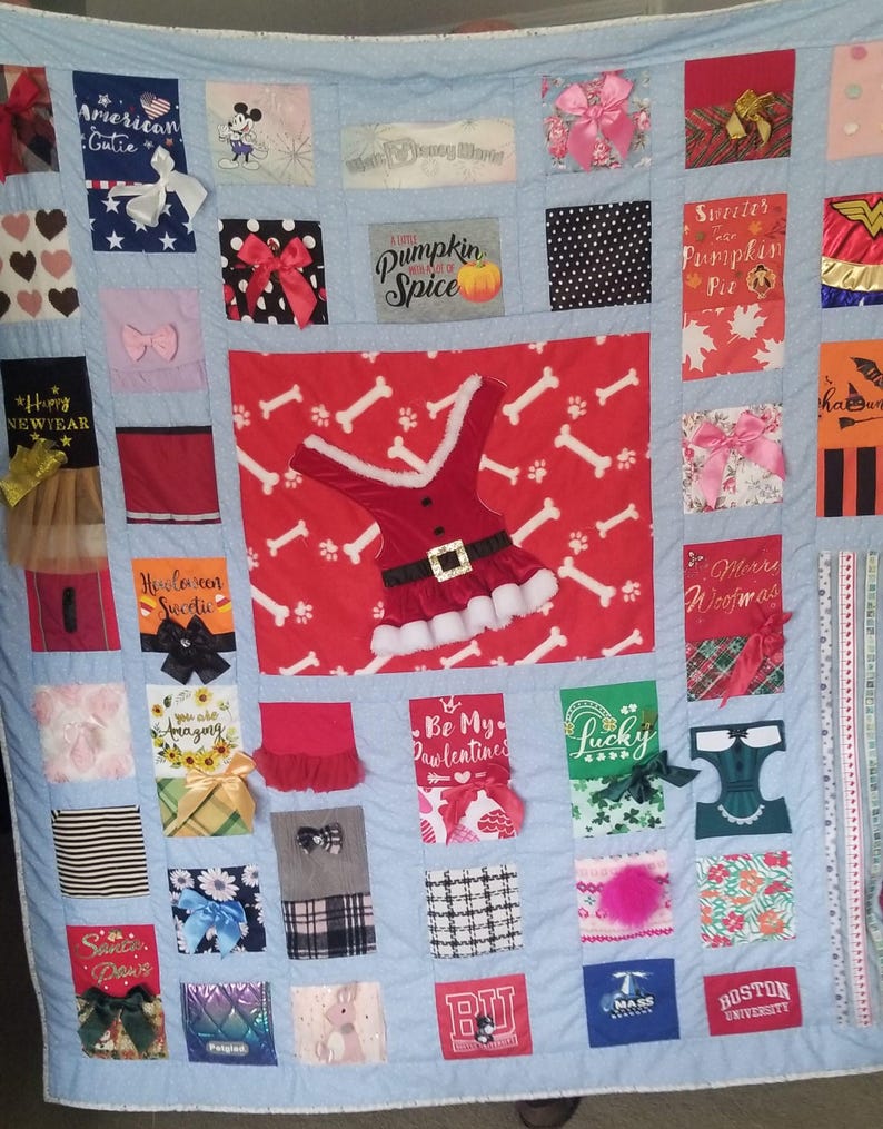Pu&ograve; includere: Una trapunta patchwork blu e bianca con vari quadrati che presentano diversi disegni e testi. Alcuni quadrati presentano testi come "Pumpkin Spice", "Be My Pawlentines", "American Gutie", "Sweater Pumpkin Pie" e "Happy New Year". Altri quadrati presentano immagini come un motivo a osso di cane, un abito di Babbo Natale, un personaggio di Topolino e un personaggio di Wonder Woman.