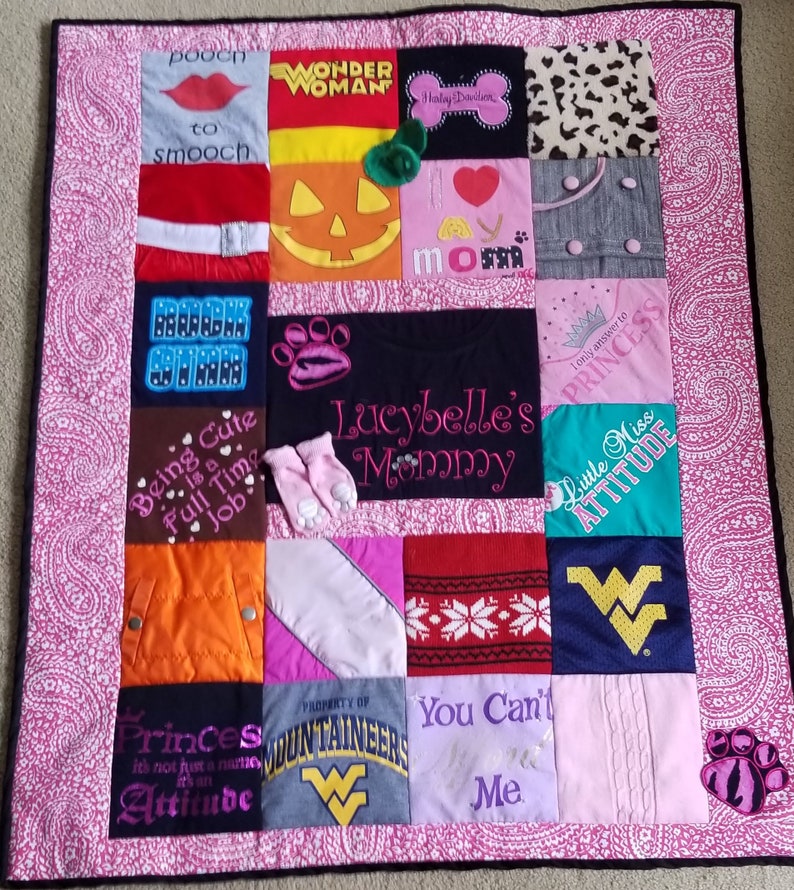 Pu&ograve; includere: Una trapunta patchwork rosa e bianca con vari quadrati che presentano diversi disegni e testi. Alcuni dei quadrati includono una zucca, un'impronta di zampa, un cuore e una corona. Il testo sui quadrati include "Wonder Woman", "Being Cute is a Full Time Job", "Princess", "Little Miss Attitude", "You Can't Me", e "Lucybelle's Mommy".